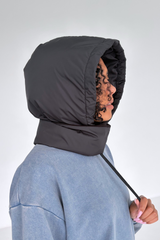A person wearing a dark gray puffer hood with a built-in neck warmer and drawstring, paired with a light blue sweatshirt, shown in profile against a light gray studio background.