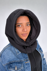 A close-up of a person wearing a black puffer-style hooded neck warmer or balaclava over a blue denim jacket, against a light gray studio background.
