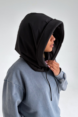 A person wearing a black hooded sweatshirt with a large attached hood over a dusty blue hoodie, shown in profile against a light gray studio background.