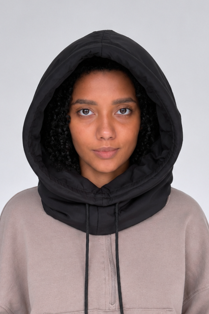 A model wearing a black hooded neck warmer or balaclava-style neck gaiter over a taupe pullover hoodie, shown against a neutral light gray background.
